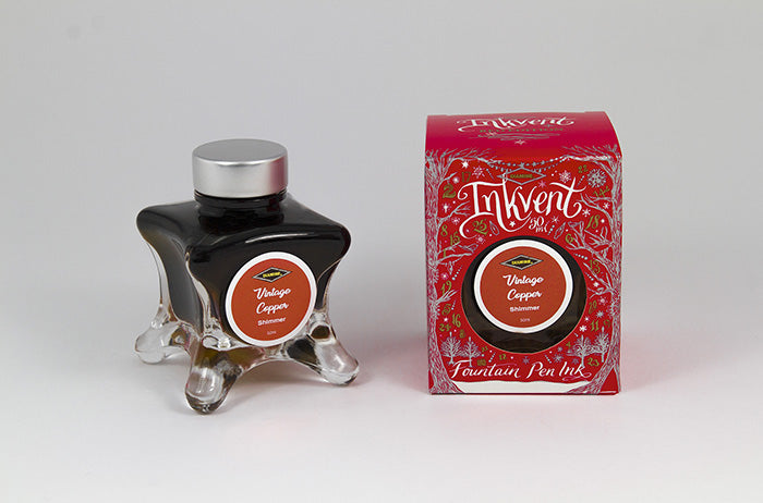 A glass bottle of 50ml Diamine Red Edition Inkvent Vintage Copper Shimmer fountain pen ink next to packaging box, in front of a white background.