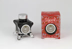 A glass bottle of 50ml Diamine Red Edition Inkvent Ash fountain pen ink next to packaging box, in front of a white background.