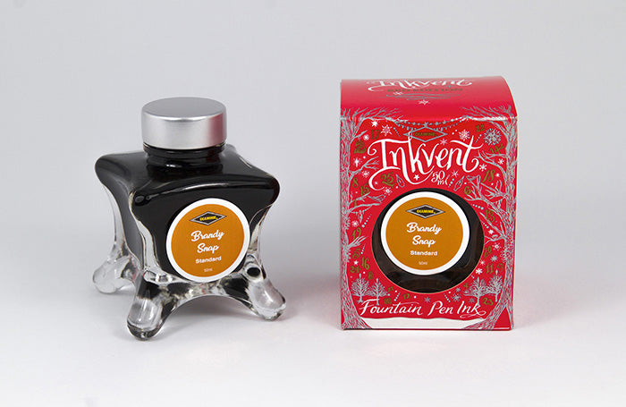 A glass bottle of 50ml Diamine Red Edition Inkvent Brandy Snap fountain pen ink next to packaging box, in front of a white background.