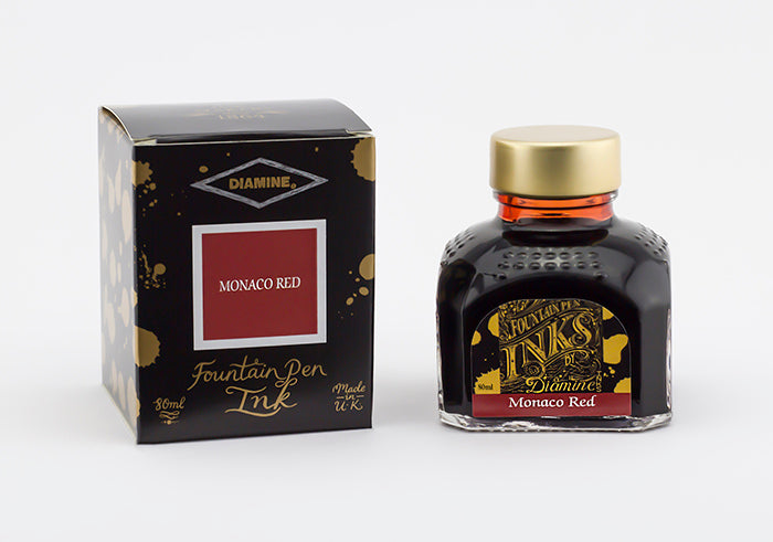 A glass bottle of 80ml Diamine Monaco Red fountain pen ink next to its packaging box, in front of a white background.