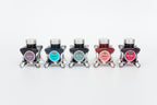Group of five glass bottles of 50ml Diamine Teal Edition Inkvent pigment fountain pen ink, Nostalgia, Let it Snow, Fir & Fog, Ambience & Ruby Taffeta. Sitting in front of a white background.