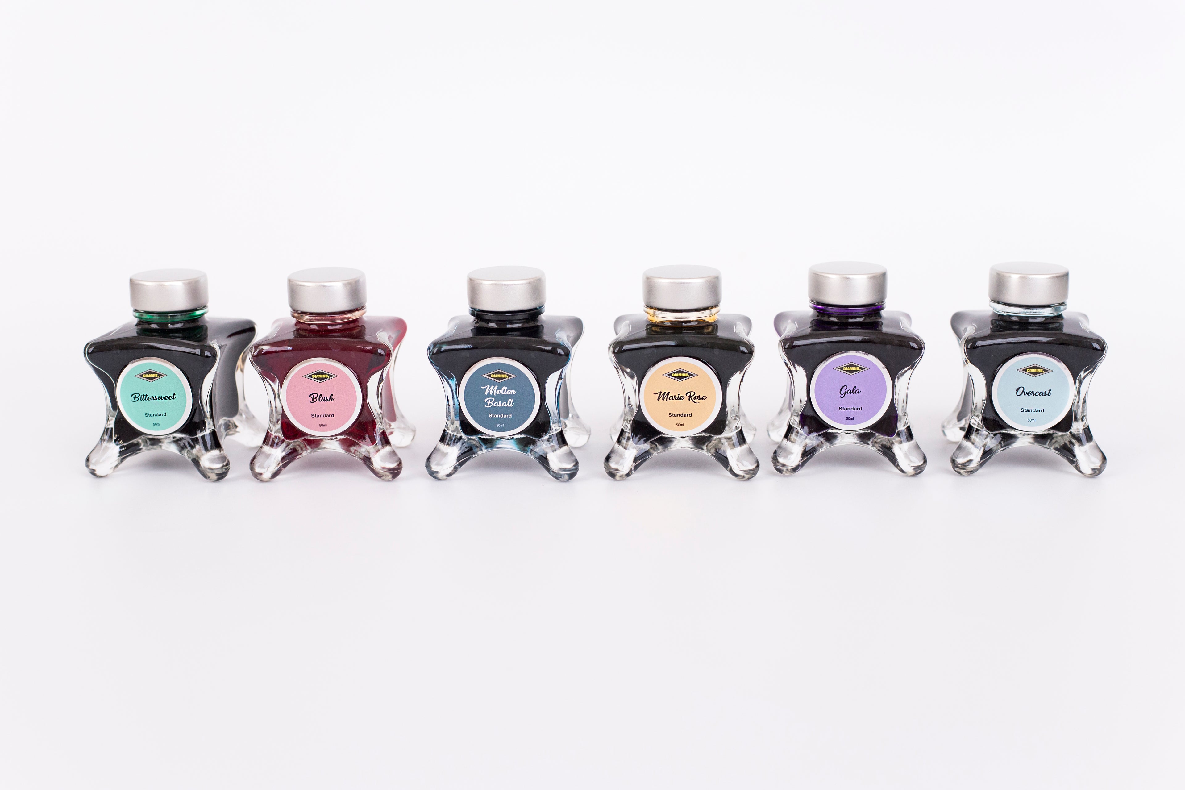 Group of six glass bottles of 50ml Diamine Teal Edition Inkvent pigment fountain pen ink, Bittersweet, Blush, Molten Basalt, Marie Rose, Gala & Overcast. Sitting in front of a white background.