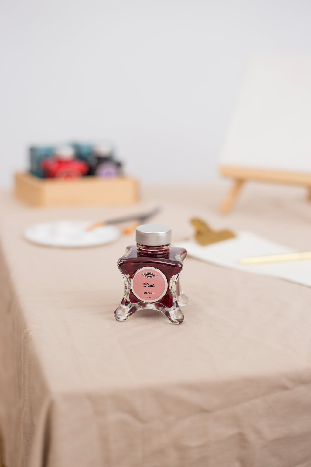 Diamine Fountain Pen Ink Bottle in Blush, on a table covered with a canvas material. Blurry background contains art supplies.