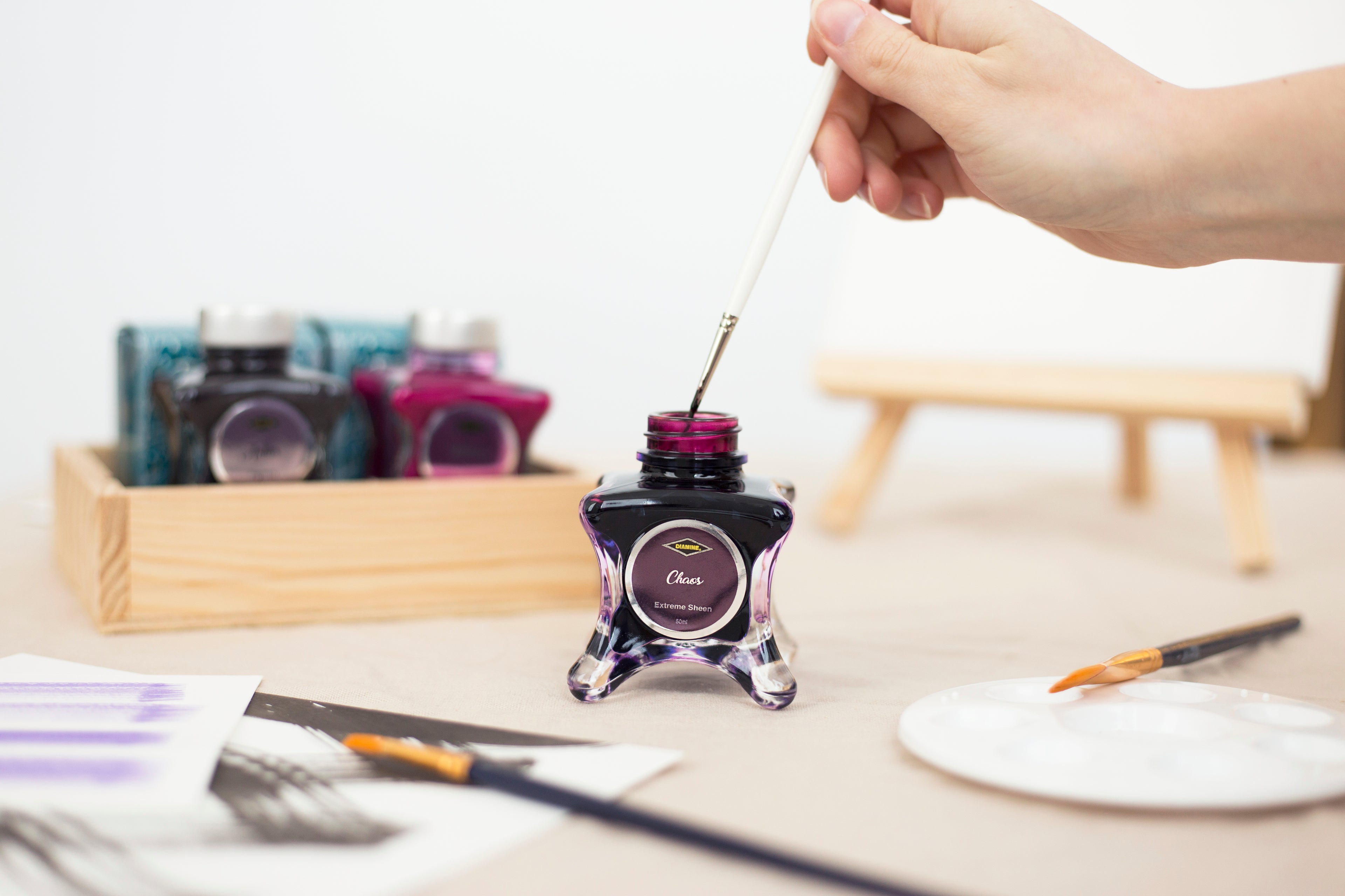 Hand dipping a paintbrush into Diamine Fountain Pen Ink Chaos, with various ink bottles and brushes on a table.