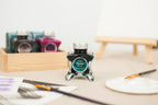 Diamine Myrrh the Merrier Fountain Pen Ink bottle with a brush and paper on a table.