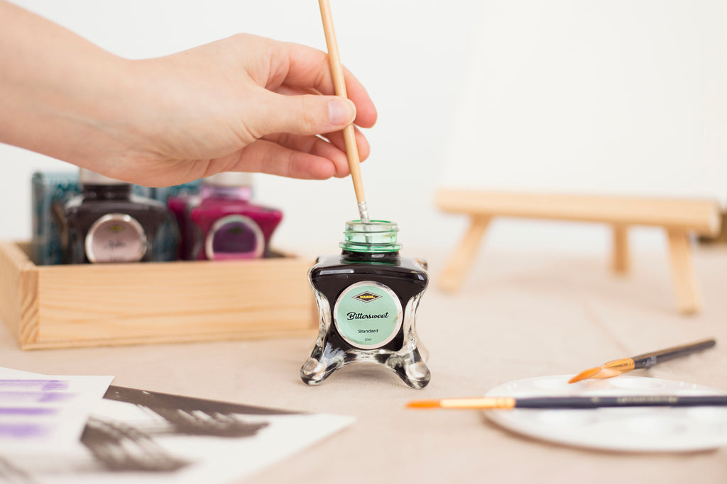 Hand dipping a paintbrush into Diamine Fountain Pen Ink Bittersweet, with various ink bottles and brushes on a table.