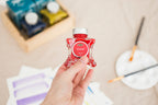Hand holding a red ink bottle (Diamine Carousel Fountain Pen Ink) with a blurred art supplies background.
