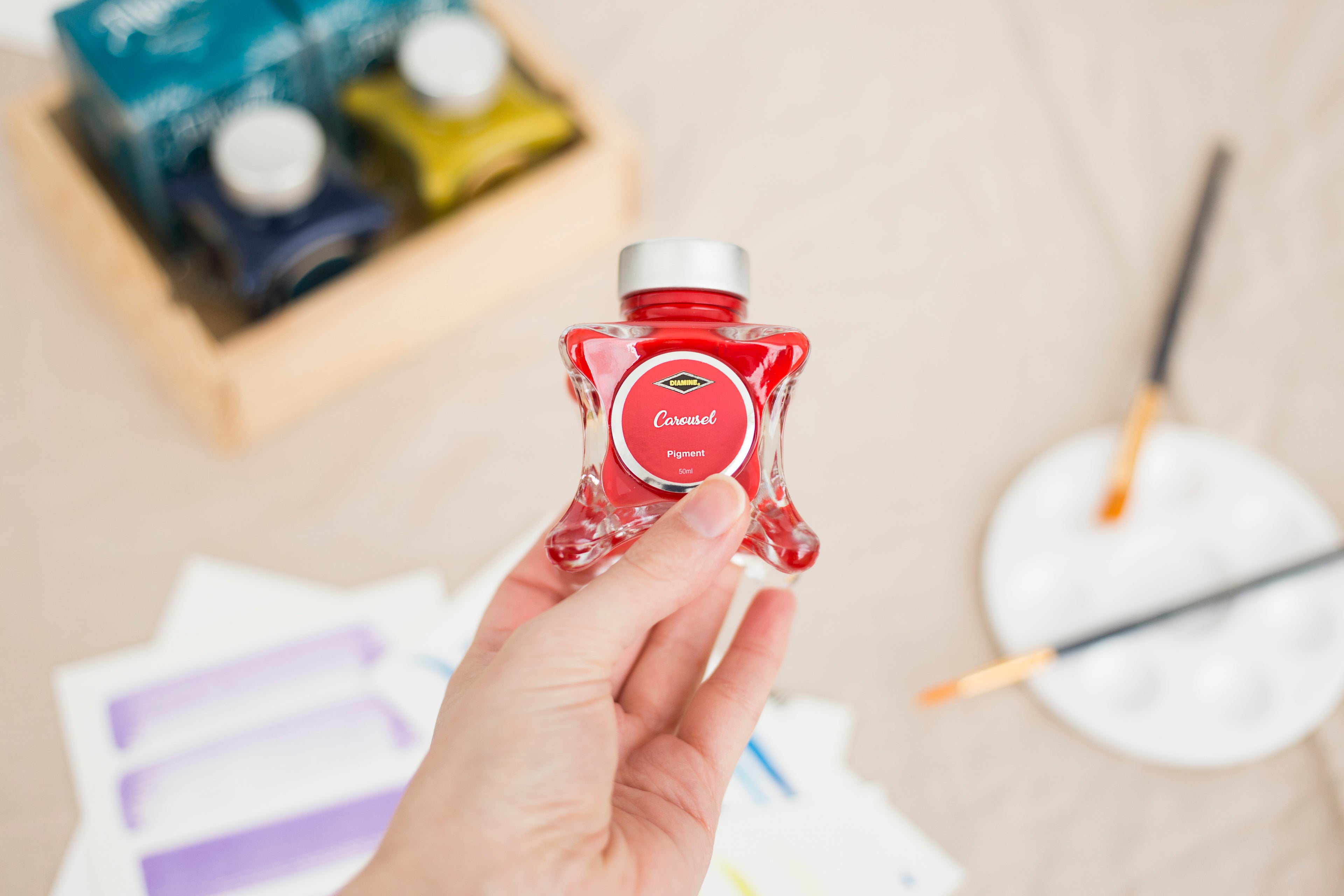 Hand holding a red ink bottle (Diamine Carousel Fountain Pen Ink) with a blurred art supplies background.