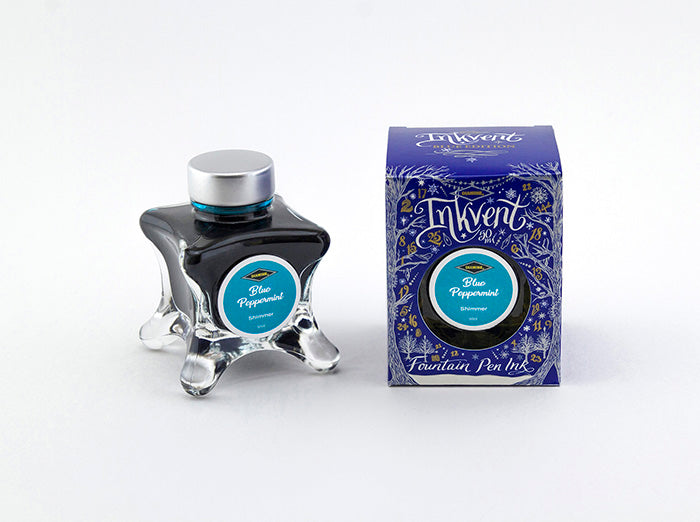 A glass bottle of 50ml Diamine Blue Edition Inkvent Blue Peppermint shimmer fountain pen ink next to packaging box, in front of a white background.